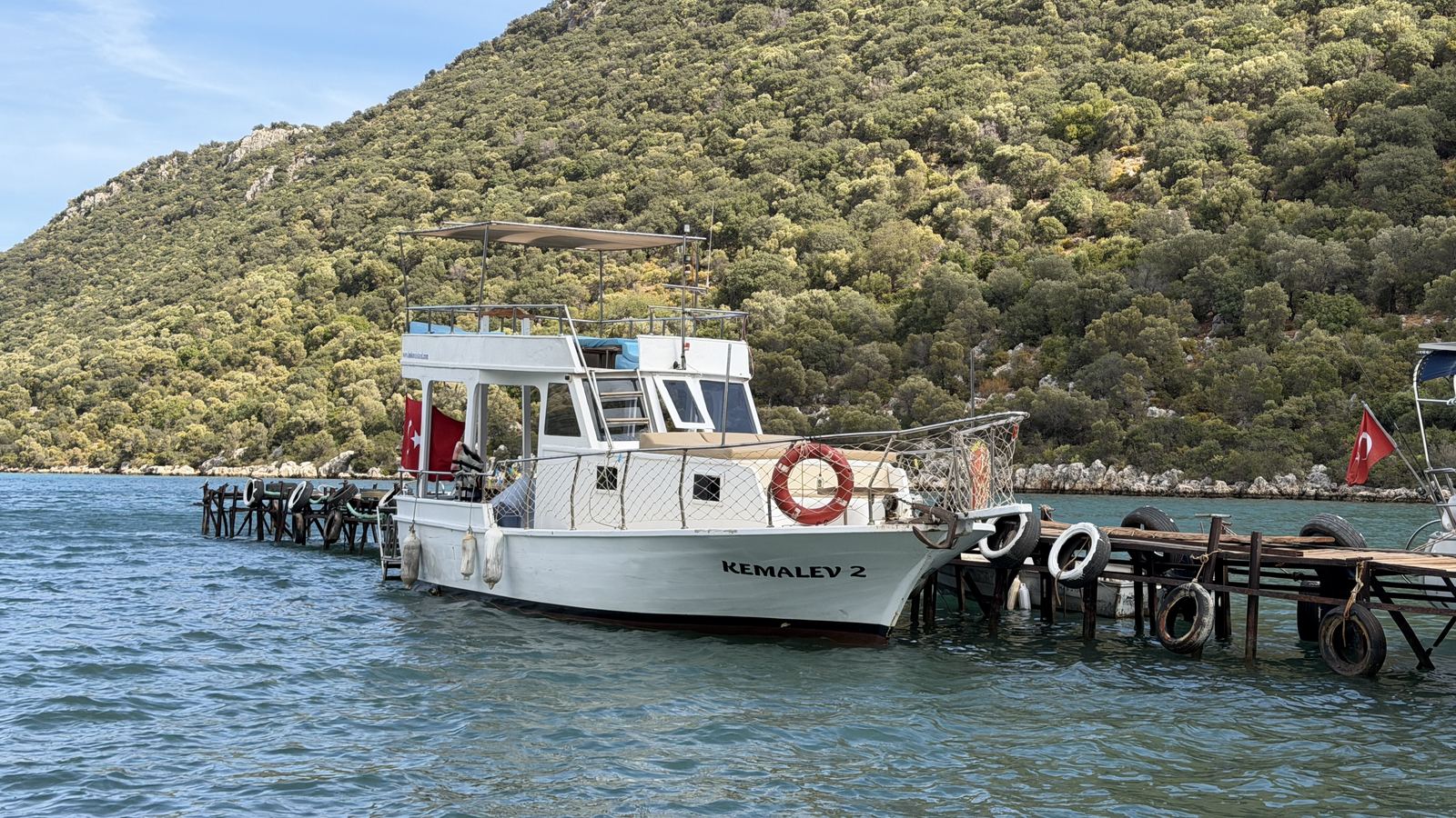 The wooden boat KEMALEV 2 moored at Üçağız pier on the Kekova strait, below a green hillside